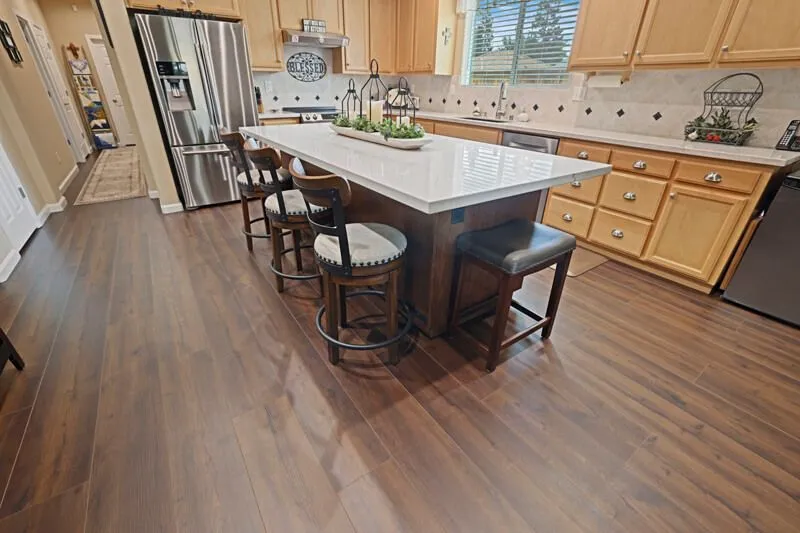 A modern kitchen with dark wood-look flooring, a large white-topped island, light wooden cabinets, and bar stools.