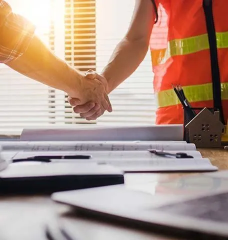 Two people in professional attire, one wearing a safety vest, shake hands over building blueprints and a model house.